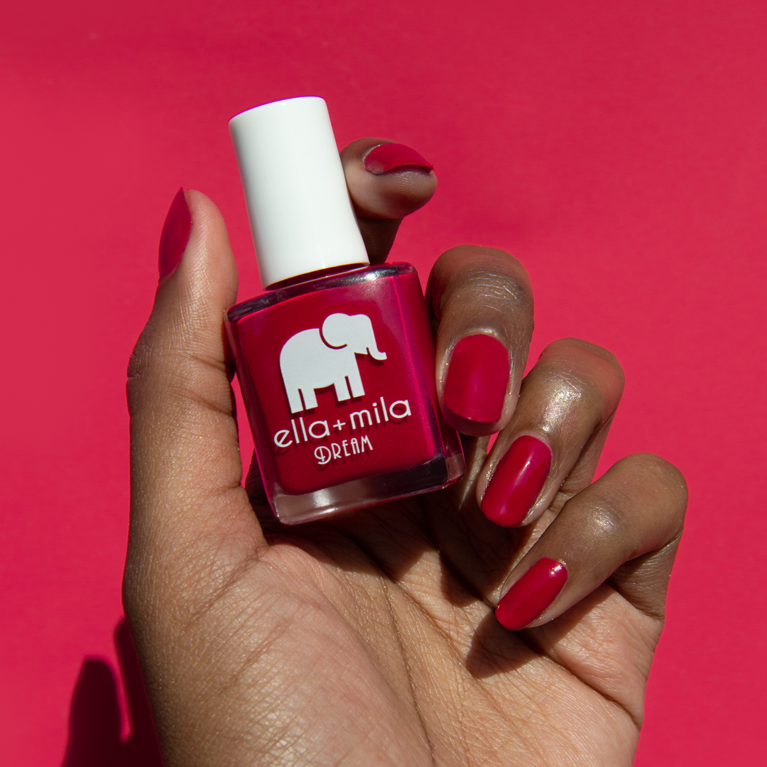Bad Obsession red nail polish on a deep skin tone against a red background, holding the bottle